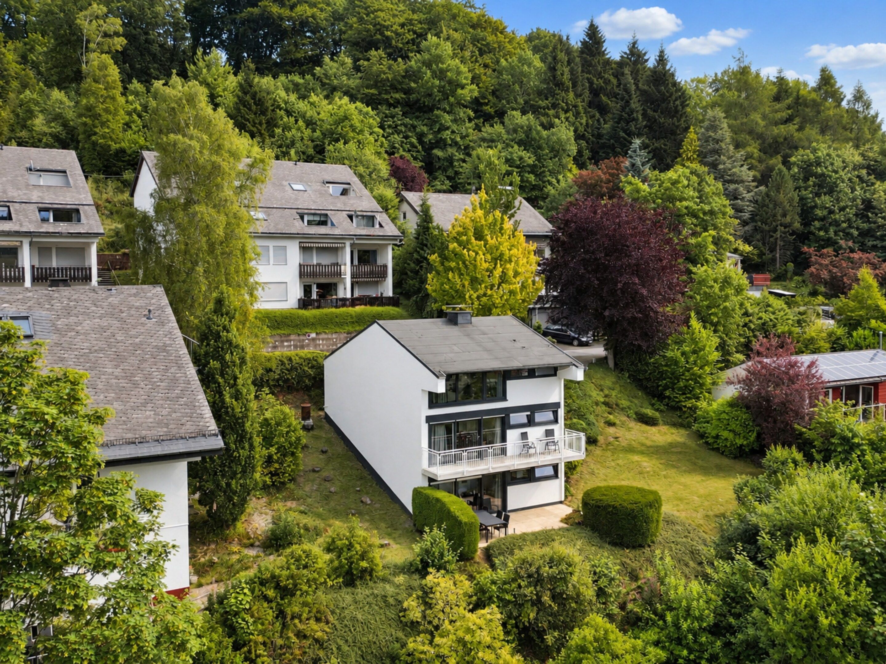 Een modern, twee verdiepingen tellend huis met een balkon, omgeven door weelderige begroeiing en bomen.