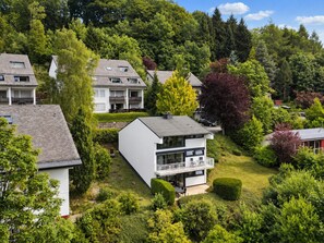 Ein modernes, zweistöckiges Haus mit Balkon, umgeben von üppiger Vegetation und Bäumen.