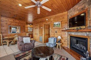 A log cabin interior with a fireplace, a television mounted above it, a ceiling fan, and a dining area with a bench and table. - 3-Bedroom Cabin w/ Game Room & Hot Tub (Gatlinburg)