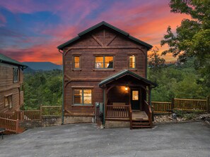 Eine zweistöckige Holzhütte mit Veranda, umgeben von Bäumen und einem Bergpanorama.
