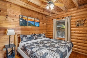 Ein Schlafzimmer in einer Blockhütte mit einem Bett, einem Deckenventilator und einem Fenster mit Vorhängen.