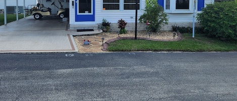 Edificio de una sola planta con puerta y ventanas azules, zona de estacionamiento techada y un pequeño jardín con una estatua de un ave.