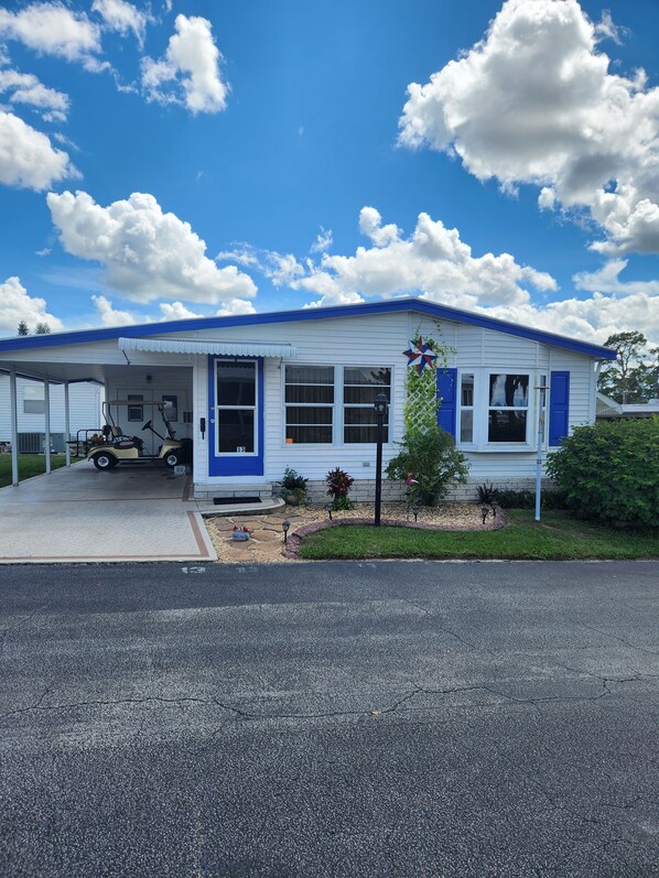 A single-story building with a blue door and windows, a covered parking area, and a small garden with a bird statue.