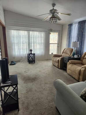 A living room with a ceiling fan, a beige sofa, a TV on a stand, and a window with curtains.