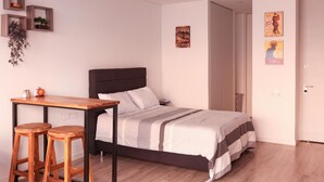A bedroom with a bed, a wooden table with two stools, a small desk, and a door. - Studio in Unicentro|Santa Barbara|Clinica Santafe (Bogotá)