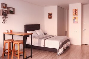 Een slaapkamer met een bed, een houten tafel met twee krukken, een klein bureau en een deur.