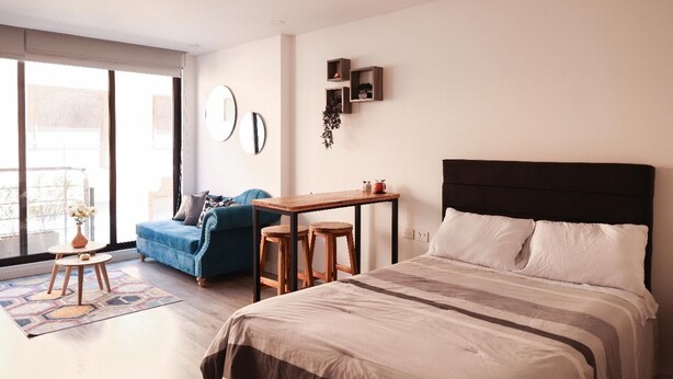 A modern bedroom with a large bed, a small table, a blue sofa, and a view through a large window. - Studio in Unicentro|Santa Barbara|Clinica Santafe (Bogotá)