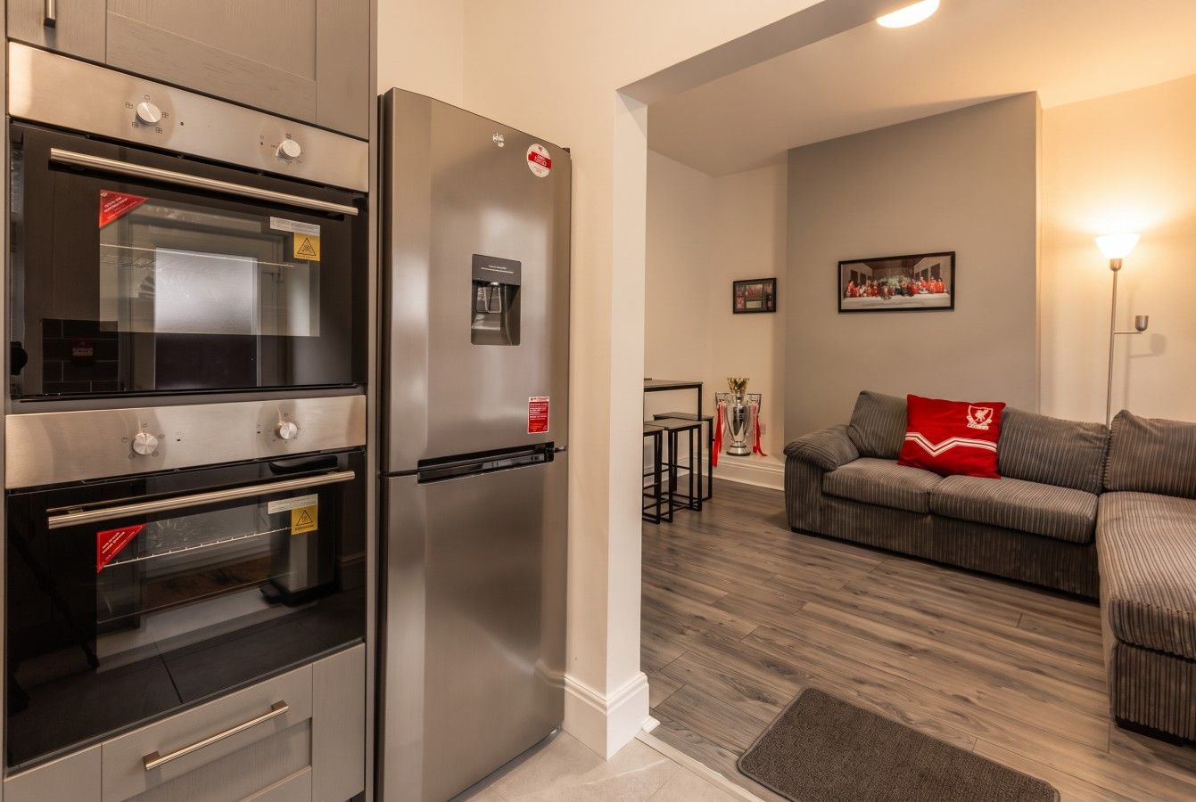 A kitchen with a built-in oven and refrigerator, a wooden floor, and a living area with a sofa and a lamp.