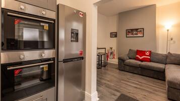 A kitchen with a built-in oven and refrigerator, a wooden floor, and a living area with a sofa and a lamp.