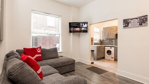 Ein Wohnzimmer mit grauem Sofagarnitur, einem an der Wand befestigten Flachbildfernseher und einer Küchenzeile mit Waschmaschine.