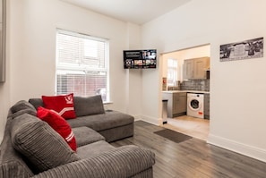 A living room with a grey sofa set, a flat-screen TV mounted on the wall, and a kitchen area with a washing machine. - En-Suite Rooms · Sleeps 8 · 5 Min Walk to Anfield Stadium (Anfield)