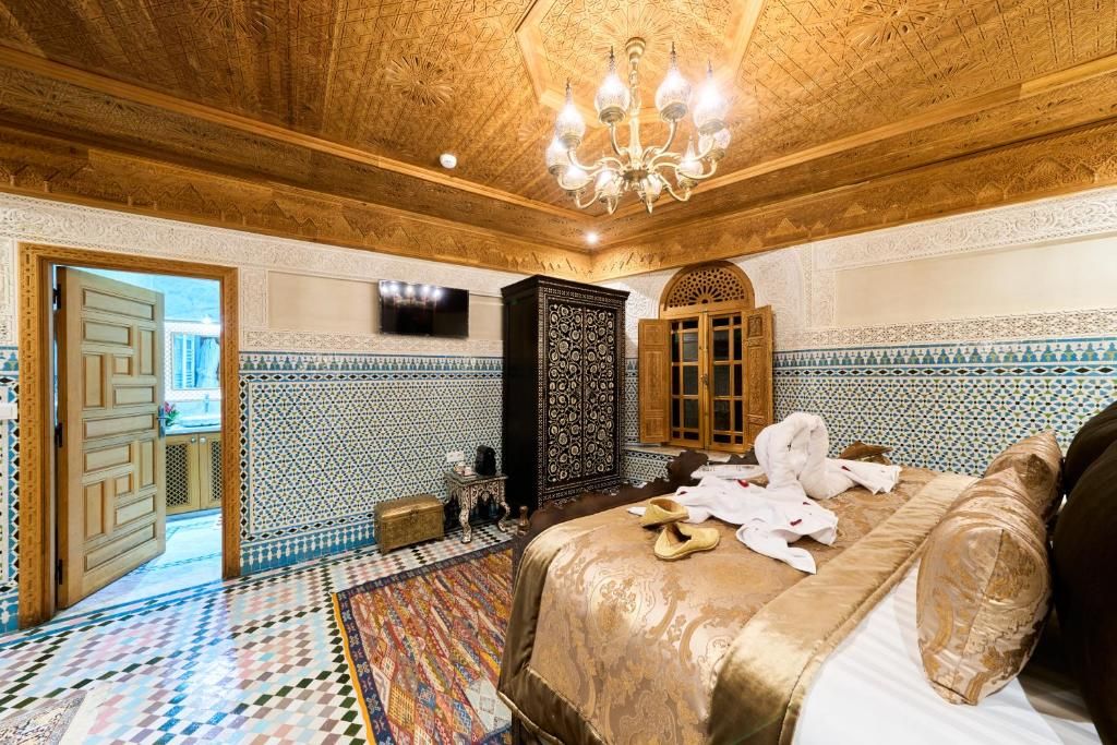 Ein Zimmer mit einem gemusterten Boden, einem Bett mit einer gemusterten Tagesdecke, einer Chandelier und einem an der Wand befestigten Fernseher.