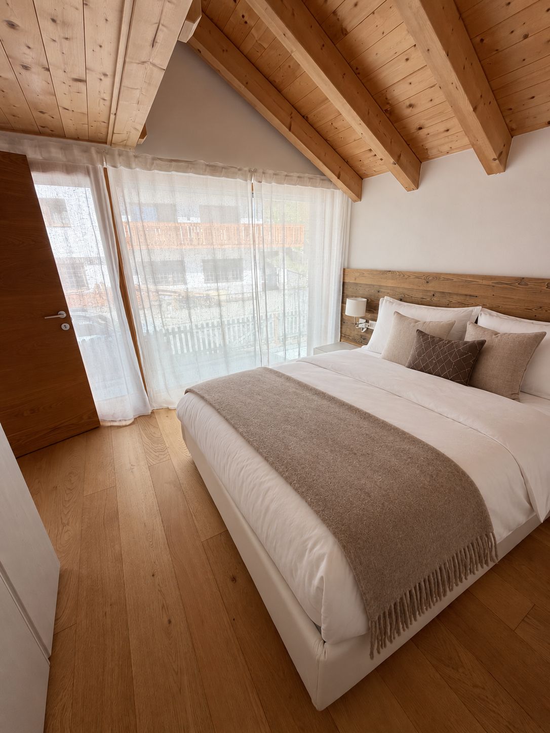 Ein Schlafzimmer mit Holzbalkendecke, einem Bett mit beiger Decke, einem Nachttisch und einem Fenster mit transparenten Vorhängen.