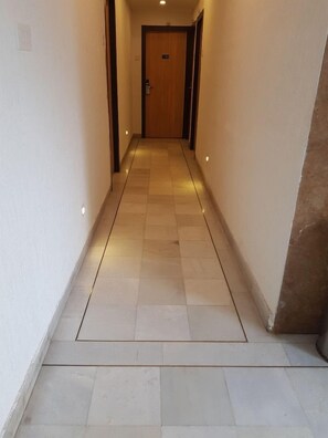 Un couloir étroit, avec une porte en bois au fond, un sol carrelé et des spots encastrés.