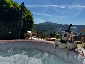 An outdoor hot tub with a bottle of wine and a glass, surrounded by a wicker chair and a table, with a mountainous landscape in the background. - La Terrazza sul Lago -lake Peace - Wonderful View (Castel Gandolfo)