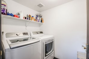 Un cuarto de lavandería con dos lavadoras y una secadora, un estante superior con productos de limpieza y una puerta a la derecha.