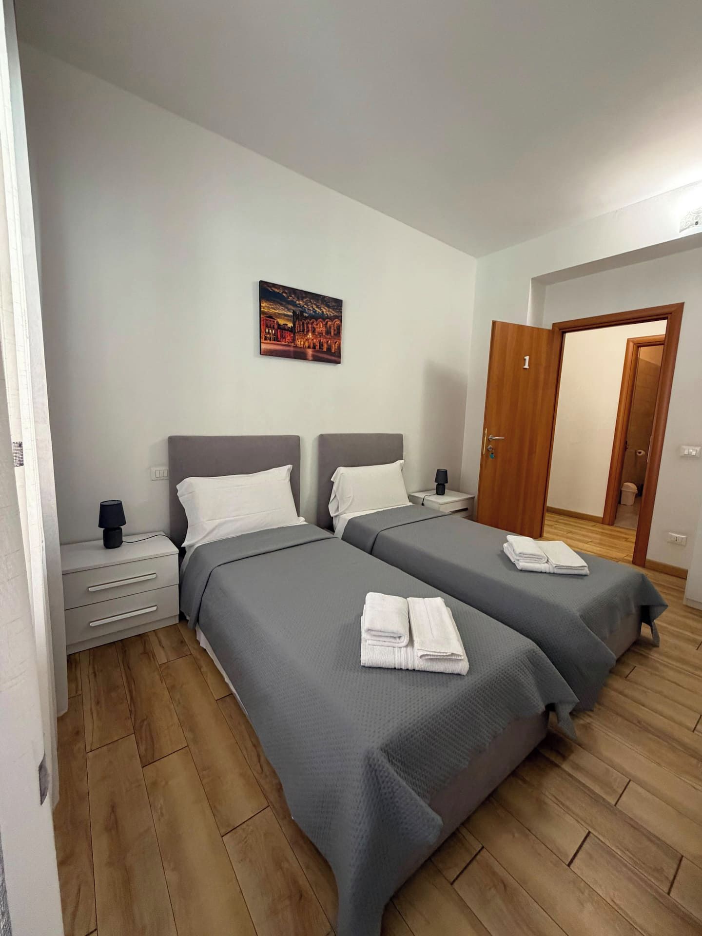 A hotel room with two beds, a nightstand with a lamp, a painting on the wall, and a wooden floor.
