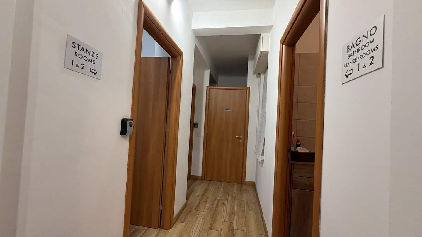 A hallway with wooden doors and signs indicating 'Stanze Rooms 1 & 2' and 'Bagno Bathroom Stanze/Rooms 1 & 2'.