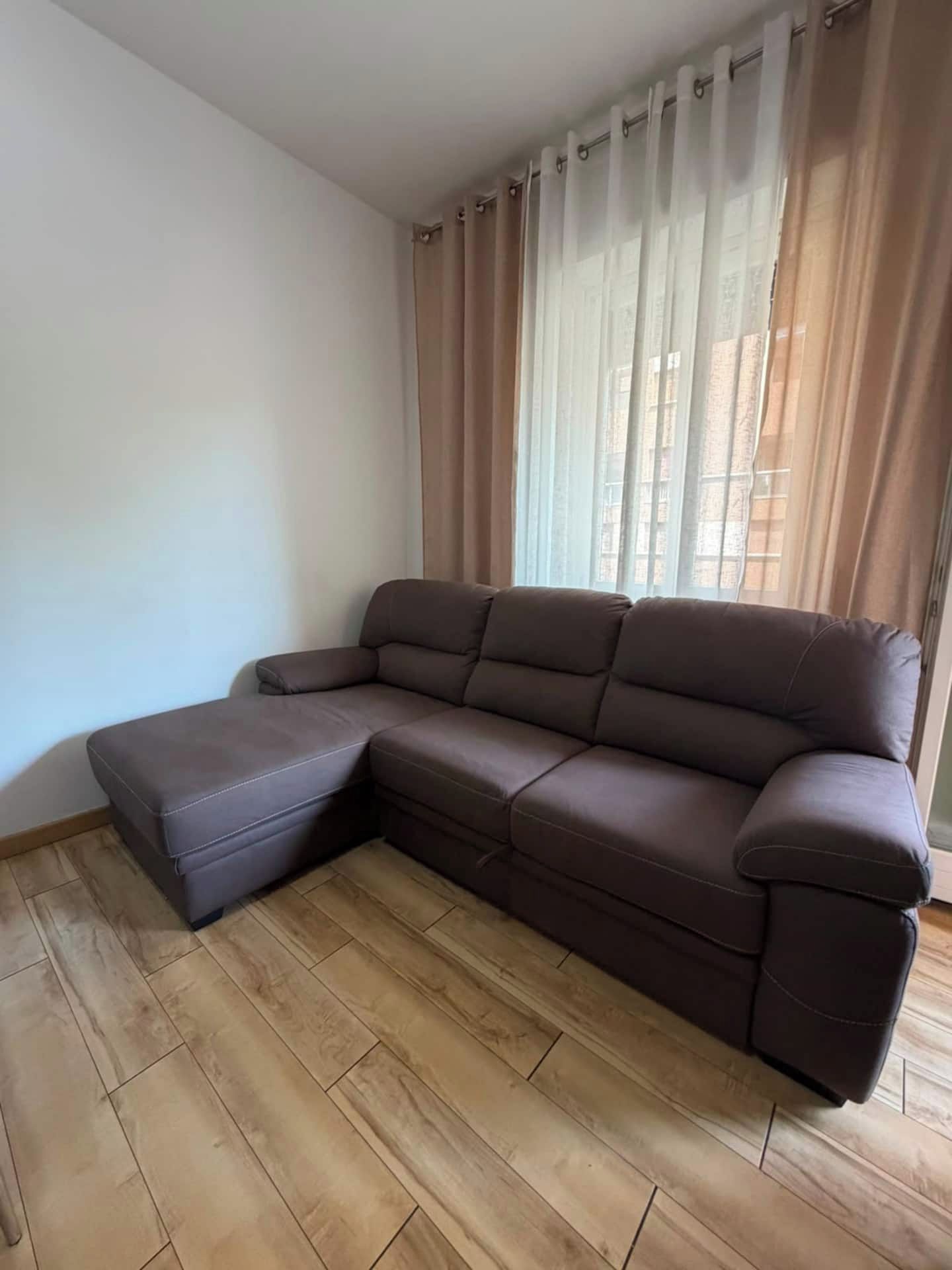 Un divano componibile con una chaise longue, posizionato su un pavimento di legno, vicino a una finestra con tende leggere.