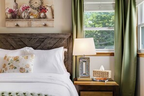 A neatly made bed with white linens and floral pillows, a bedside lamp, a small bedside table with a tissue box and books, and a window with green curtains.