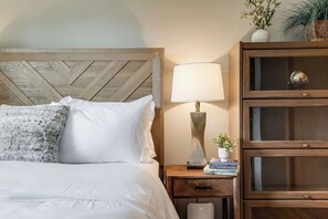 A neatly made bed with white linens and a fluffy pillow, a bedside table with a lamp and a small plant, and a wooden cabinet with glass doors.