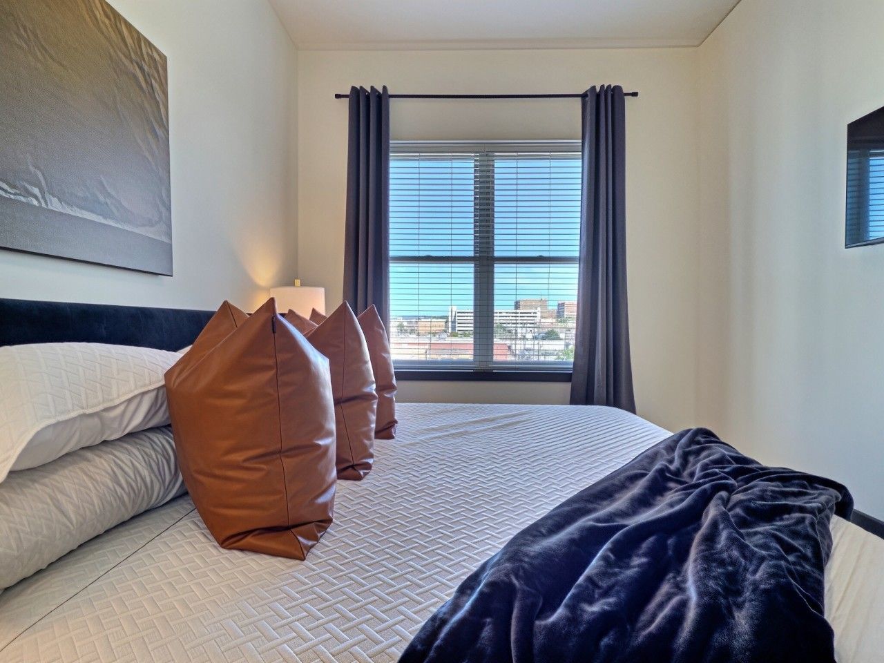 A hotel room with a bed, pillows, a blanket, a window with curtains, and a painting on the wall.