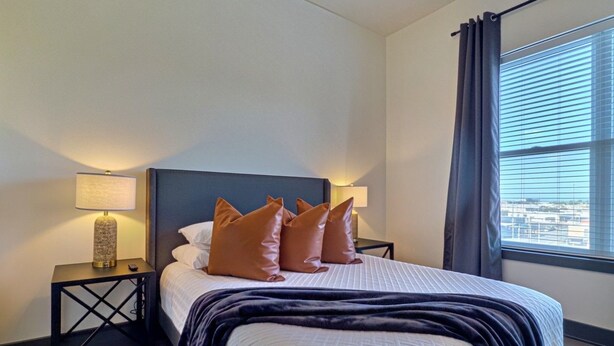 A hotel room with a bed, two bedside tables, a lamp, a window with curtains, and a view of the city. - Two bedroom two bath, walk to UAB and dining (Birmingham)