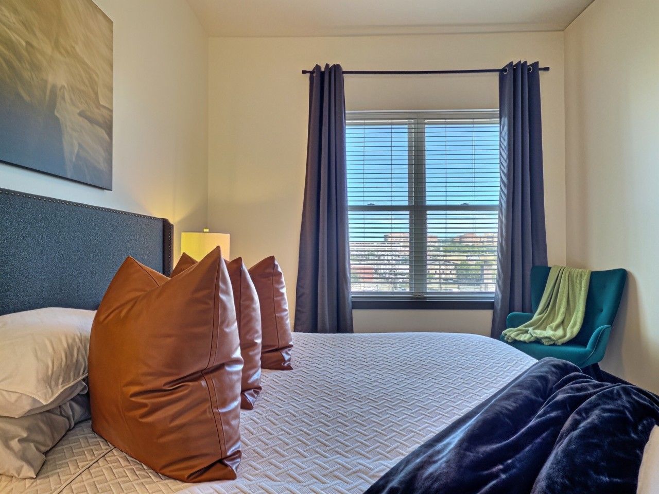 Habitación de hotel con cama, almohadas, ventana con cortinas, una silla y un cuadro en la pared.