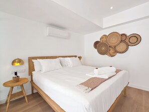 Cama bem arrumada com roupa de cama branca, mesa de cabeceira com abajur e ar-condicionado fixado na parede.