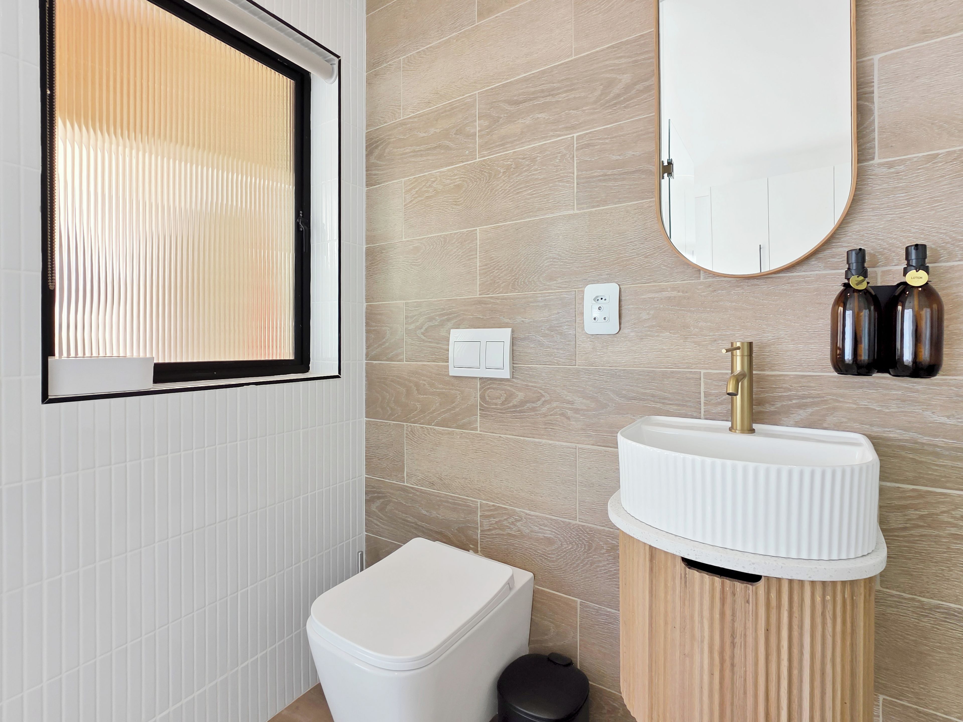 A bathroom with a white toilet, a wooden vanity with a white sink, a mirror, and two bottles of lotion on the wall.