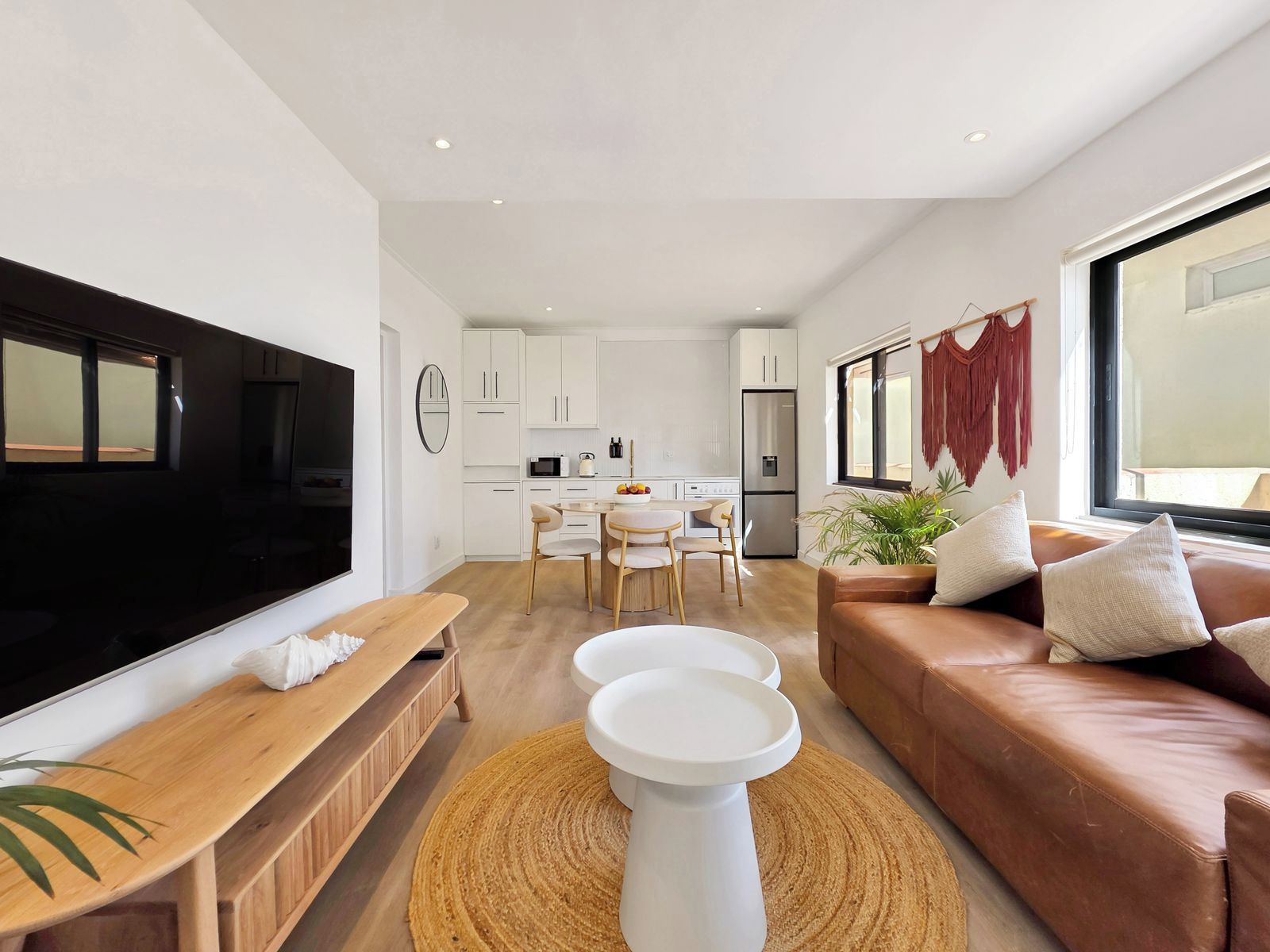 Una sala de estar moderna con un televisor de pantalla plana, un sofá de cuero marrón, un banco de madera y un comedor con una mesa y sillas blancas.