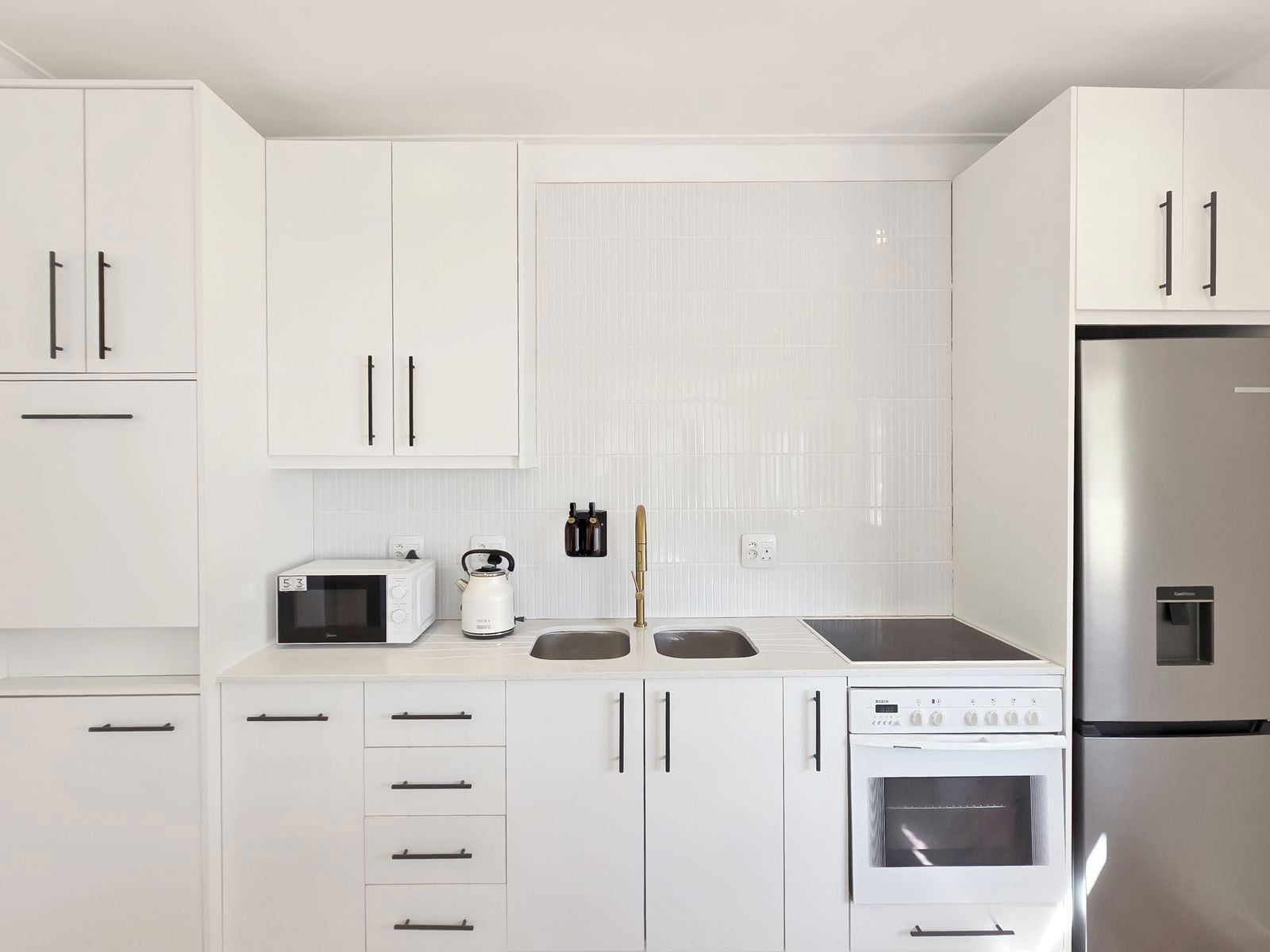 A modern kitchen with white cabinets, a Bosch oven, a microwave, and a stainless steel refrigerator.