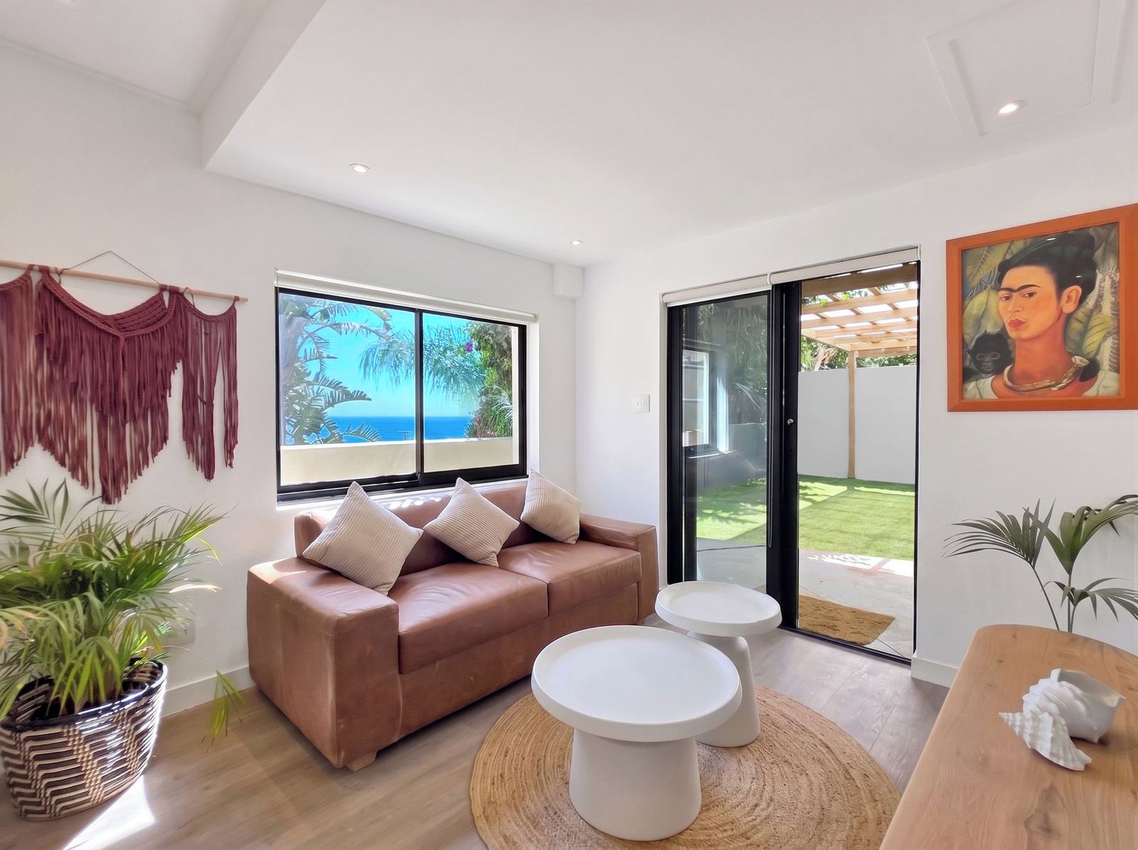 A modern living room with a sofa, two side tables, a macrame wall hanging, and a large window with a view.