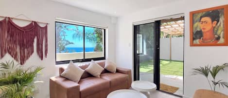A modern living room with a sofa, two side tables, a macrame wall hanging, and a large window with a view.