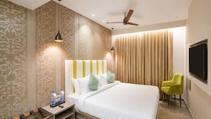 A modern hotel room with a large bed, a bedside table with water bottles, a chair, a TV, and a ceiling fan. - Deluxe the Beacon (Visakhapatnam)