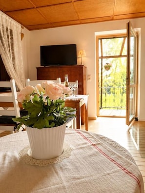 Ein Zimmer mit Esstisch für zwei Personen, einer weißen Vase mit Blumen, einem Fernseher an der Wand und Blick ins Freie durch eine Glastür.