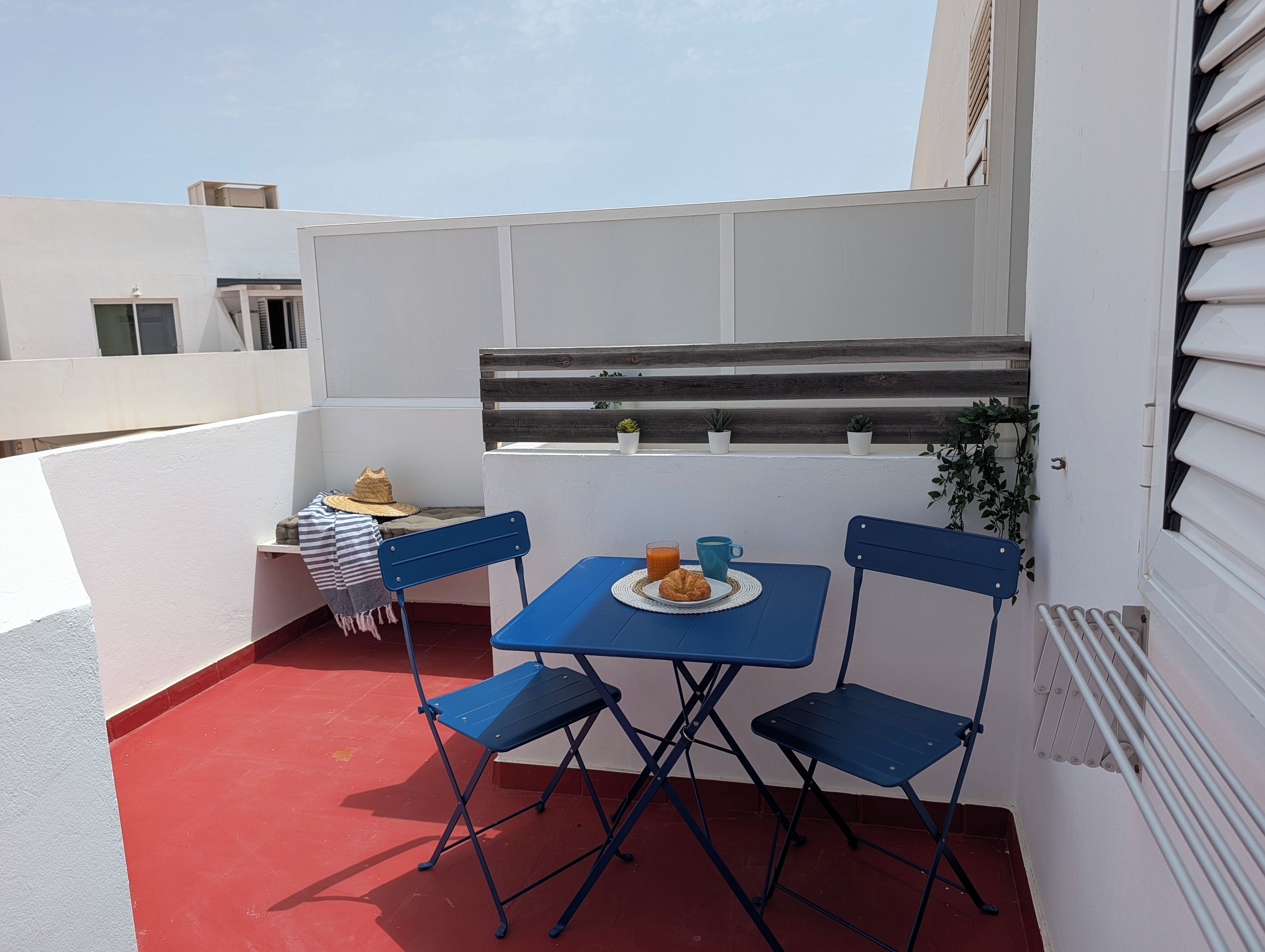 A small outdoor seating area with a table and two chairs, a potted plant, and a window with shutters.