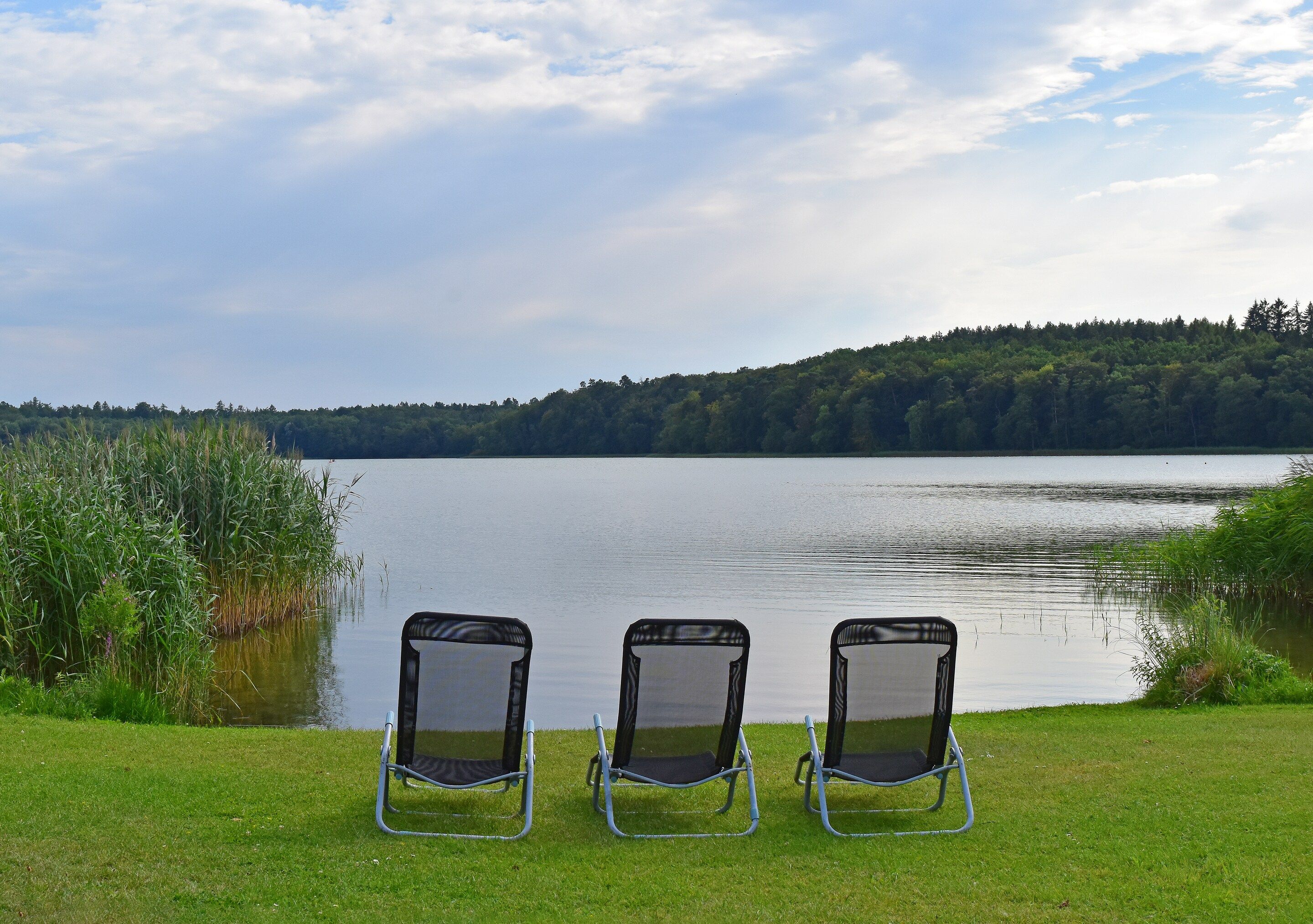 Three outdoor chairs facing a lake with a forested hill in the background.