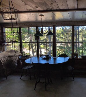 A dining area with a round table, chairs, hanging lights, and a view of trees through large windows.