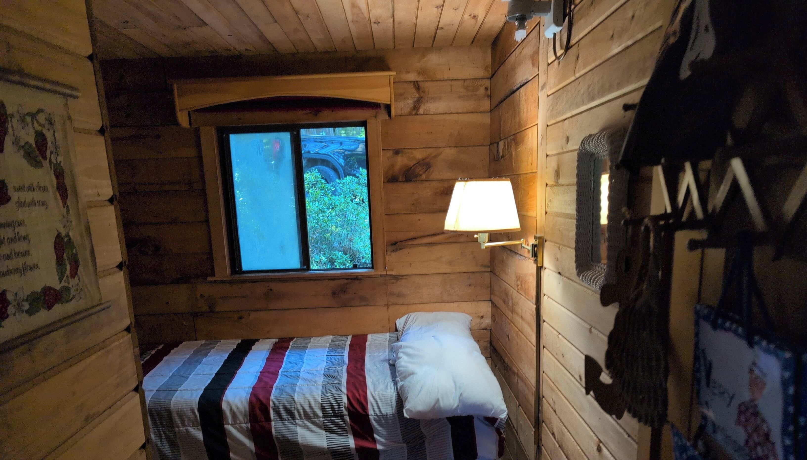 Ein kleiner Holzraum mit einem Einzelbett, einem Fenster mit Blick ins Grüne und einer Wandlampe.