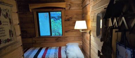 Ein kleiner Holzraum mit einem Einzelbett, einem Fenster mit Blick ins Grüne und einer Wandlampe.