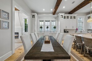 Un comedor con una larga mesa de madera, sillas blancas y una cocina al fondo.