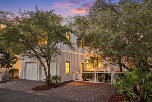 Una casa moderna de dos pisos con garaje, rodeada de árboles maduros y un camino de entrada pavimentado.