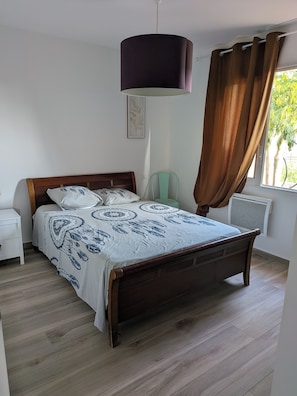 A bedroom with a wooden bed, a bedside table, a nightstand, a hanging lamp, a window with curtains, and a radiator. - House with a pool and hot tub just a stone's throw from the Canal du Midi. (Saint-Nazaire-d'Aude)