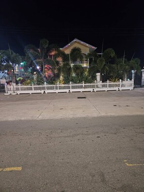 Vista nocturna de un edificio con valla blanca al frente, rodeado de palmeras y un área pavimentada.