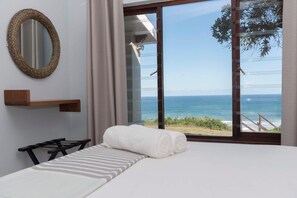 A hotel room with a bed, a wooden shelf, a round mirror, and a large sliding glass door leading to a balcony with a view of the sea. - Brenton Breakers (Knysna)