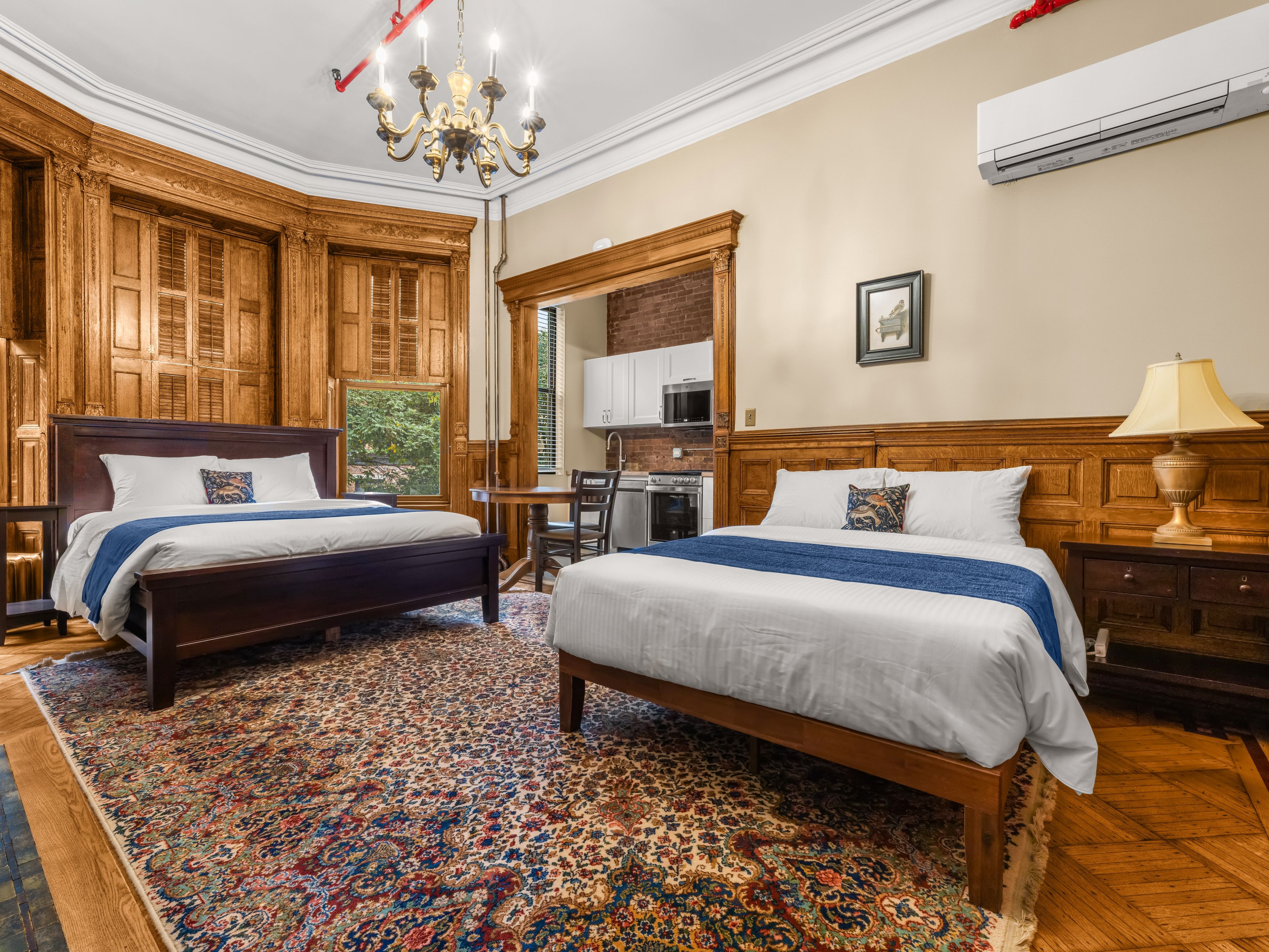 Habitación de hotel con dos camas, un candelabro, cabecero de madera, alfombra estampada y una cocina visible a través de una ventana.