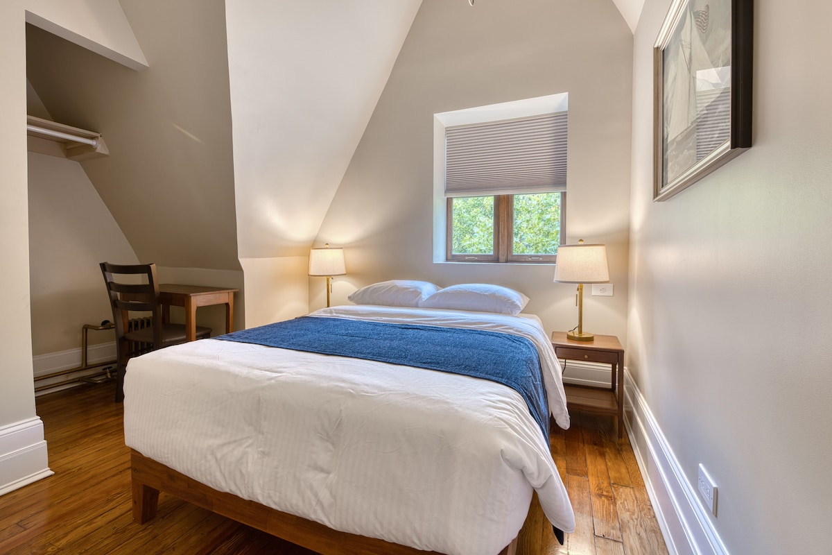 A bedroom with a sloped ceiling, a bed with white bedding and a blue throw, two bedside lamps, a window with blinds, and a chair.