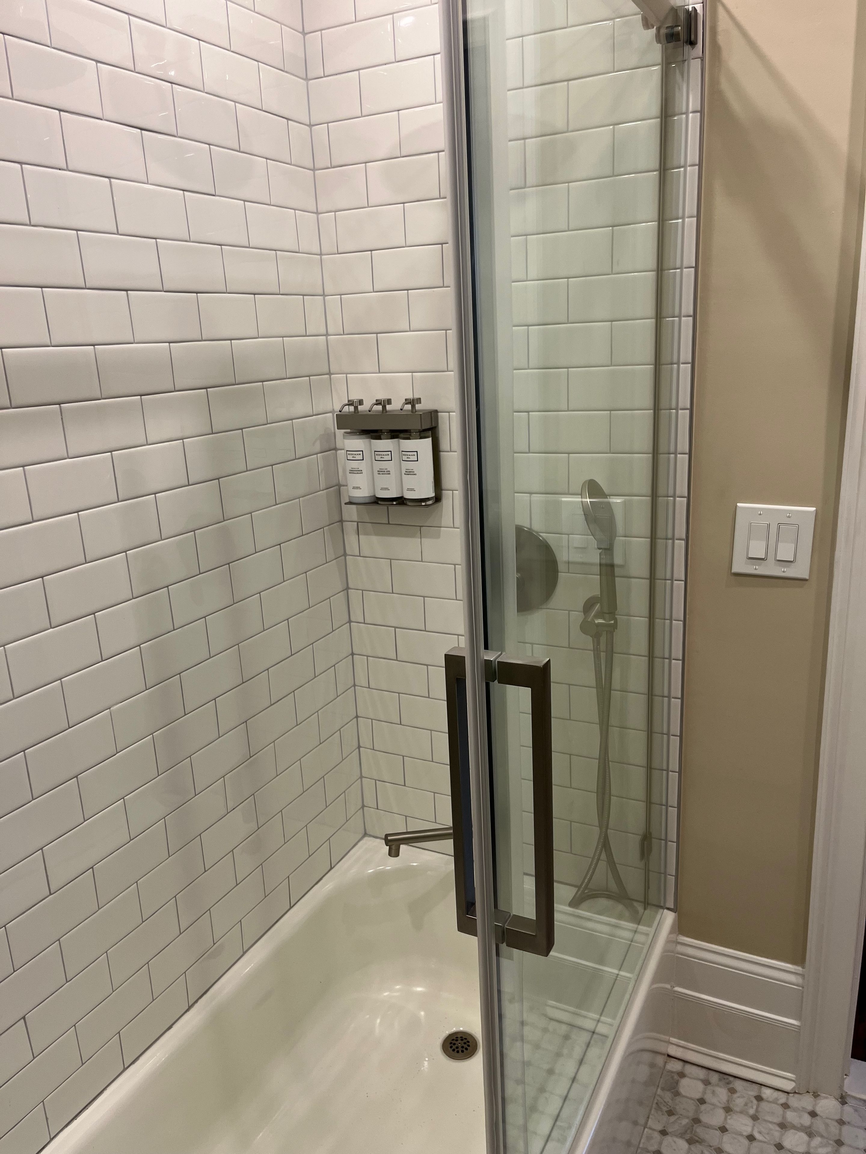 Un espace douche doté d’une porte en verre, de murs recouverts de carreaux blancs et d’un pommeau de douche.