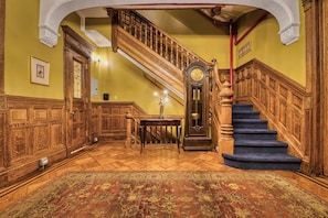 A grand staircase with a wooden handrail and blue carpeted steps, leading to an upper floor. A large, ornate clock is mounted on the wall above the stairs.